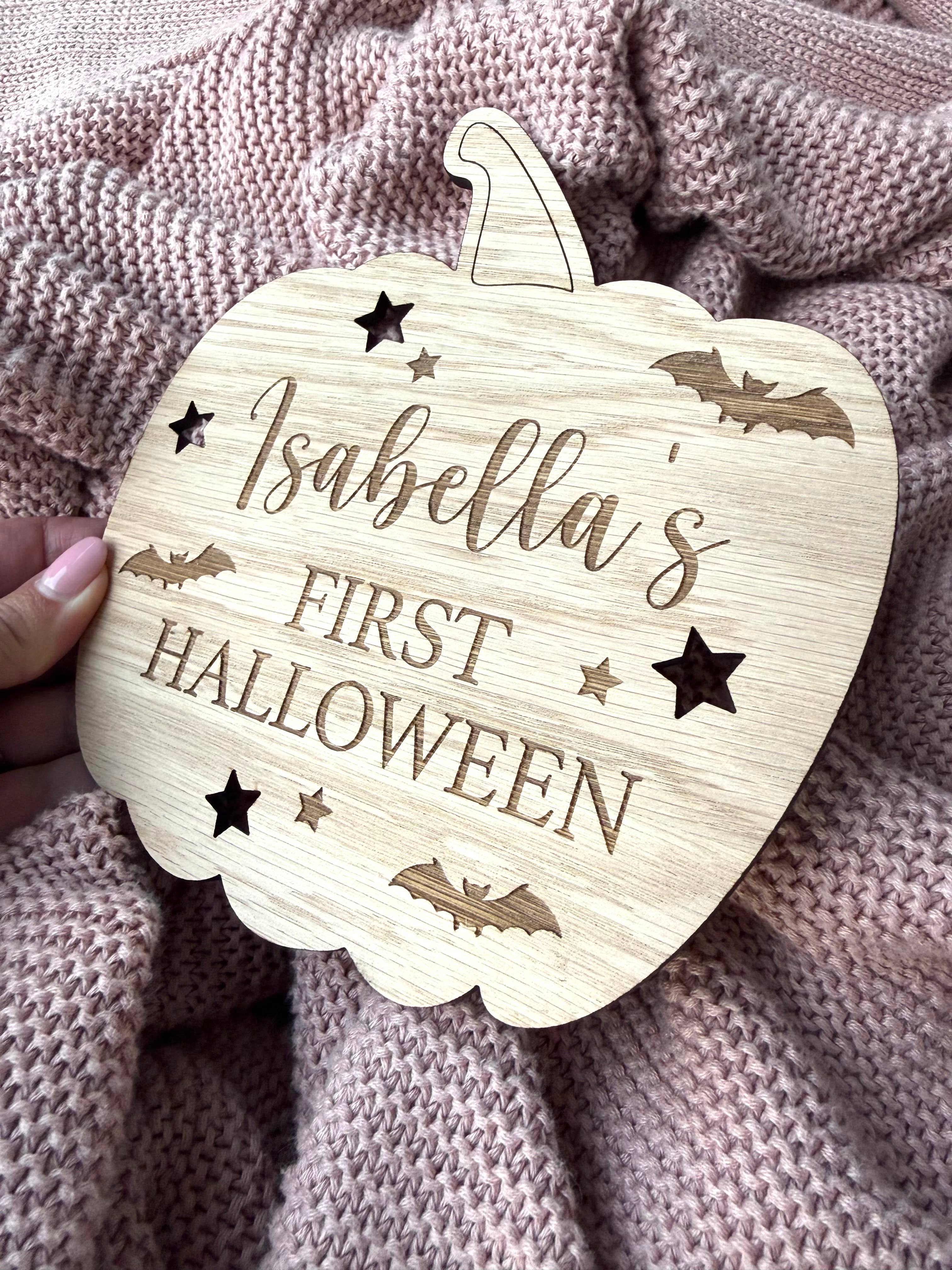 Wooden pumpkin-shaped ornament with 'Isabella's First Halloween' text on a textured fabric background